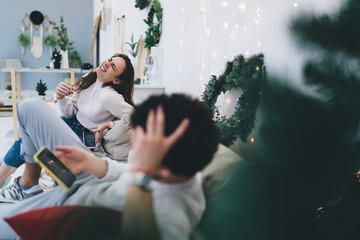 Fototapeta na wymiar Cheerful caucasian woman laughing at funny content from social networks resting at festive apartment with husband, couple in love using technology for blogging and searching during winter vacations