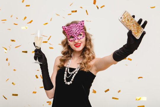young stylish woman on white background drinking champagne, making self photo, celebrating new year, wearing black dress, fashion jewelry, mask, happy carnival disco party, holding glass, having fun