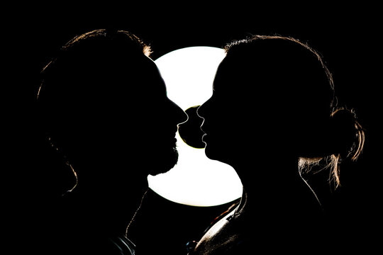 Couple of man and woman looking closely at each other and kissing in the dark illuminated by a light behind them.