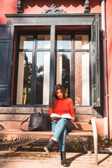 Fototapeta na wymiar Lifestyle. Young Caucasian brunette in a red shirt sitting on a bench in the park reading a book. Vertical photo
