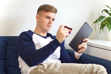 Confused worried young man holding a computer, tablet and credit bank blocked card in hands and having problems with paying, buying online. Internet fraud