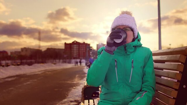 Female person sits on wooden bench drinks cocoa from disposable mug in public park at sunset in winter. Woman finishes her coffee from paper cup turns it over slaps bottom checking remains of drink
