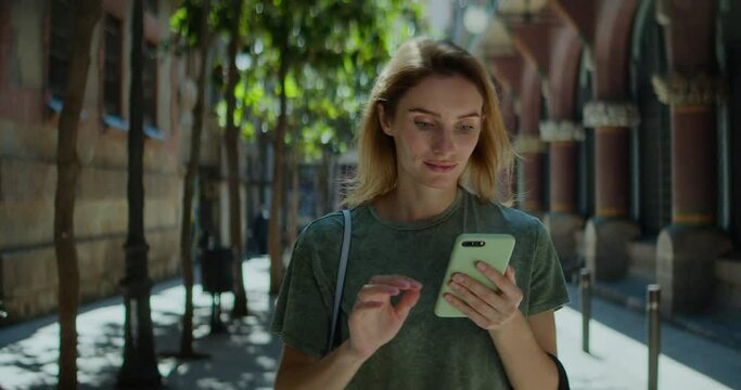 Young Pretty Woman Typing on Smartphone While Walking Streets of Summer City. Backward Tracking Shot of Attractive Girl Using Mobile Device for Communication or Navigation. Slow Motion Cinematic