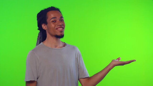 Product advertisement. Serious strict young man presents copy space on green background croma key, indicates with index finger, shows place for commercial text or goods, wears gray t shirt