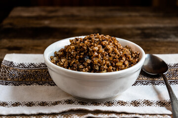 Buckwheat porridge in a white ceramic bowl, metal horse, kitchen napkin on a wooden table. Homemade food preparation.