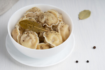 Pelmeni or dumplings of Russian cuisine made of minced meat filling wrapped in dough served in white bowl with black pepper seasoning and bay or laurel leaf on white wooden background. Horizontal