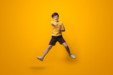 Fototapeta na wymiar Jumping caucasian boy with red hair is wearing glasses and point at camera wearing a yellow shirt on a studio wall