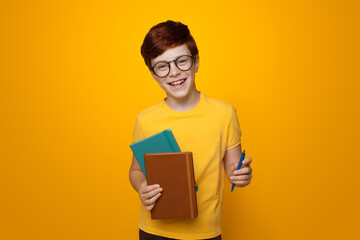 Young ginger schoolboy holding some folders is smiling on a yellow studio wall while wearing glasses and casual t-shirt