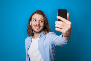 Young long haired man with beard is smiling and making a selfie using a phone on a blue studio wall in casual clothes