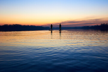Silhouettes of two women paddle on stand up paddle boarding (SUP) on quiet winter or autumn Danube river at sunset. Colorful sunset over the river with silhouettes of people