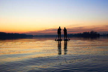 Silhouettes of two women paddle on stand up paddle boarding (SUP) on quiet winter or autumn Danube...