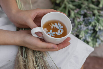 Cup of tea in female hands of a young girl outdoor, with blurred flowers on background. Mug with tea and blue flowers inside. .