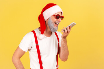 Smiling happy grey bearded man in santa claus hat and stylish glasses holding smartphone near mouth talking to voice assistant, leaving voice message. Indoor studio shot isolated on yellow background