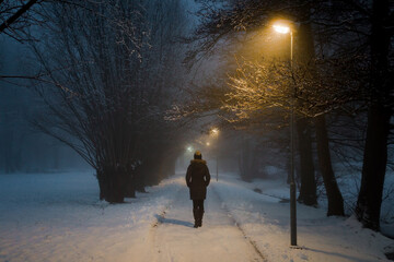Young adult woman alone slowly walking on sidewalk under yellow street lights in mist. Foggy air....