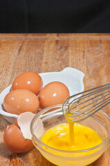 Top view, medium distance of a raw, scrambles egg in glass bowel with whisk, egg shells and three brown eggs in ceramic, white bowel, on a wood cutting board and black background