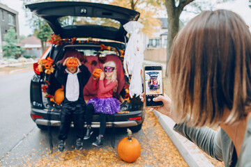 Trick or trunk. Children boy and girl with red pumpkins celebrating October Halloween holiday in trunk of car outdoors. Mother taking pictures of kids on smartphone camera for social media.