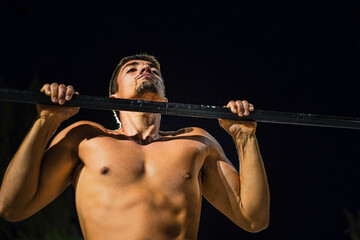 Young shirtless guy training at night street workout. Doing pull-ups.