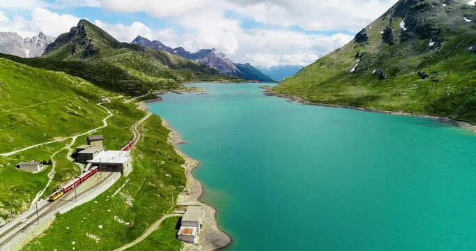 Red train passing near a turquoise blue lake in Switzerland at the bottom of a mountain. Aerial view with a drone of the beautiful landscape,under the blue sky 4K