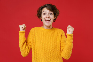 Excited happy joyful young brunette woman 20s in basic casual yellow sweater standing clenching fists doing winner gesture looking camera isolated on bright red colour background studio portrait.