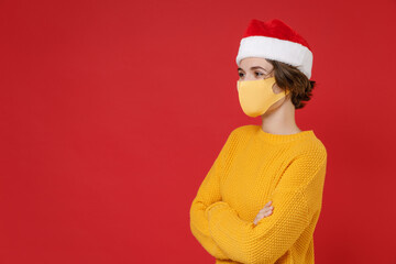 Side view of young Santa woman in Christmas hat face mask safe from coronavirus virus covid-19 hold hands crossed isolated on red background studio. Happy New Year celebration merry holiday concept.