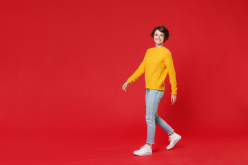 Fototapeta na wymiar Full length side view of smiling cheerful beautiful attractive young brunette woman 20s in basic yellow sweater walking going looking camera isolated on bright red colour background studio portrait.