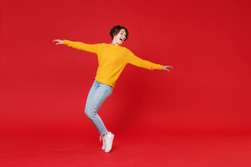 Full length side view of funny attractive young brunette woman 20s wearing basic yellow sweater dancing standing on toes spreading hands isolated on bright red colour background studio portrait.