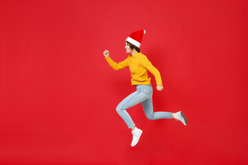 Fototapeta na wymiar Full length side view of smiling young Santa woman in yellow sweater Christmas hat jumping like running isolated on red background studio portrait. Happy New Year celebration merry holiday concept.