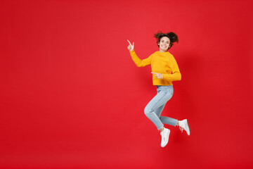 Full length side view of smiling young brunette woman 20s in basic yellow sweater jumping pointing index fingers aside on mock up copy space isolated on bright red colour background studio portrait.