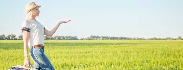Sexy blonde girl in a hat with a suitcase on a background of green field, with space for text. Banner