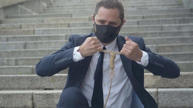 Portrait of frustrated businessman in Covid-19 face mask checking suicidal rope. Young depressed Caucasian man attempting to commit suicide. Coronavirus impact.