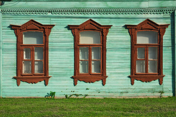 Three windows in a row on a wall of an old russian house decorated with wood carving and painted in aqua and brown colors. Traditional russian style architecture.