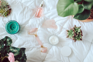 Facial beauty roller, cream, serum and green plants on white bed. Morning beauty routine concept