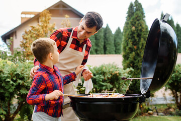 Young father and his son are cooking barbecue on an outdoor grill in their home backyard and having...