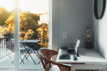 An interior of a cozy room with a desktop and a laptop on it with a USB hub and wireless graphic tablet near on the table; a glass door leads to a small balcony with a beautiful sunny view and a table - obrazy, fototapety, plakaty