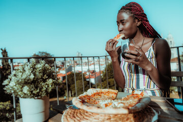 Portrait of a young charming pensive black woman with braided hair having lunch while sitting on a balcony of an outdoor pizzeria, biting a piece of delicious vegan pizza and drinking red wine