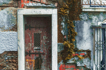 Texture of an old flaked stone facade wall of an antique house with a doorway, red bricks, stones, and cracked plaster, old dark-red door with wrought iron hinges and a lattice in the center