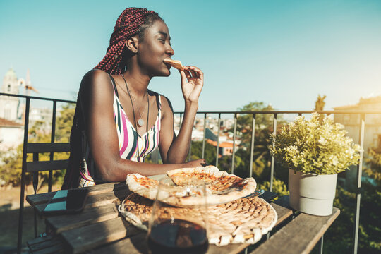 Portrait Of A Hungry Young Black Woman Biting A Piece Of Margherita Pizza During Her Brunch While Sitting At The Table Of An Outdoors Restaurant Located On The High Floor, A Glass Of Wine Near Her