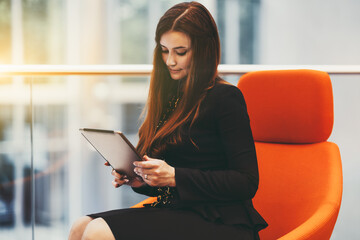 Portrait of a charming mature caucasian woman entrepreneur in a black strict costume having a group video call using her digital tablet while sitting in an office open-space area on an orange armchair