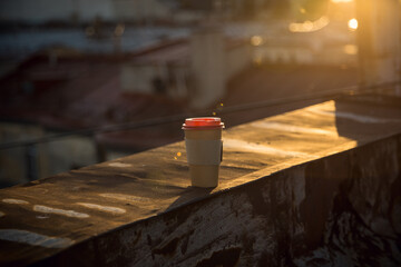 A disposable cup of coffee with a red lid stands on the rusty roof of the city at sunset. View of...