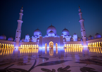 Amazing Sheikh Zayed Grand Mosque at night time, illuminated and crescent visible between two minarets. People visit the mosque for praying. Muslim mosque concept.