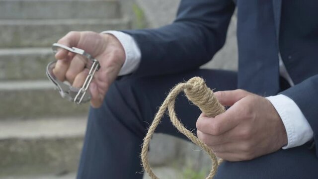 Unrecognizable man holding handcuffs and suicidal rope in hands. Desperate criminal businessman choosing between imprisonment and death.