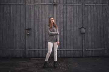 Fototapeta na wymiar beautiful young blonde model girl. White pants. gray knitted sweater. black boots. wooden pendant on the neck in the form of a horse. On the Sunset. on a background of gray wooden gates