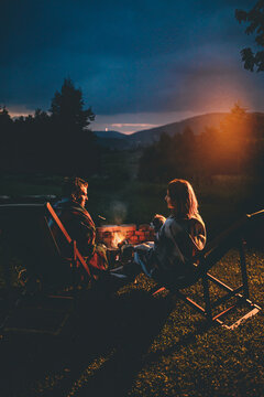 Romantic Couple With Cute Dog Relaxing In Campsite With Fire Pit. Burning Campfire With Mountain Landscape With Night Sky Over The Forest And Hills. Getaway In Wild Nature.