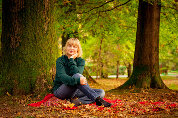 a beautiful middle-aged woman,blonde,sitting under a large tree,on a red blanket of autumn leaves,on an autumn Sunny day