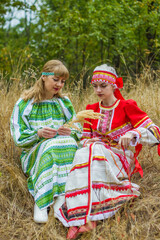 Two young women of different ages in folk costumes of red and green, sitting in the grass, chatting, playing.