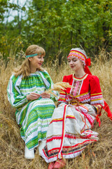 Two young women of different ages in folk costumes of red and green, sitting in the grass, chatting, playing.