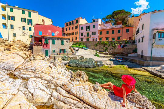 Woman in red dress sitting on cliffs at Marciana Marina in Borgo al Cotone with the famous houses of old village in bay of Marciana Marina promenade, Elba Island, Tuscany