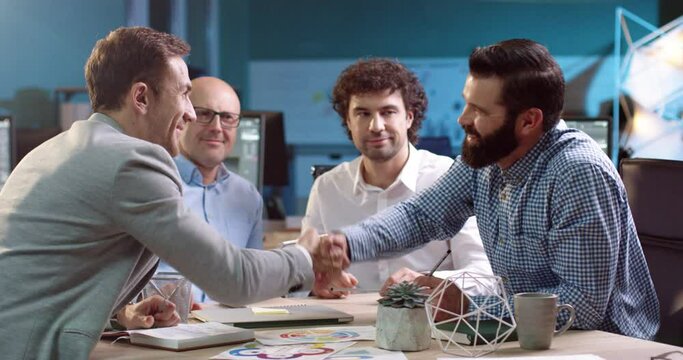 Group of business analysts sitting at table with documents. Two adult men sign agreement by shaking hands.
