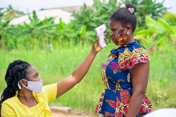 African health worker testing a lady's temperature-young black women with face mask-Black nurse with thermometer gun during a Covid-19 pandemic.