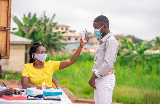image of African health worker in face mask checking temperature during covid-19 season-black church usher pointing thermometer gun on a church member before entry with Bible and records book on table